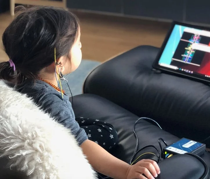 A young girl receiving neurofeedback on a black couch in her home, watching a colorful graph on the neurofeedback device.