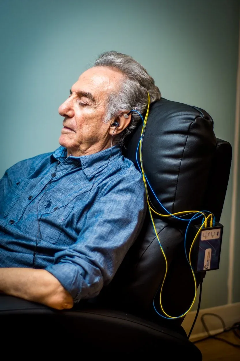 An elderly man sitting in a black chair with his eyes closed receiving neurofeedback session device attached to the side of the chair, with yellow and blue sensors. He appears to be relaxing with his head tilted back.