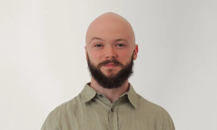 A headshot of owner, Nigel Barnim, bald headed, full beard, and mustache, wearing a beige button-up shirt, standing against a plain white background.
