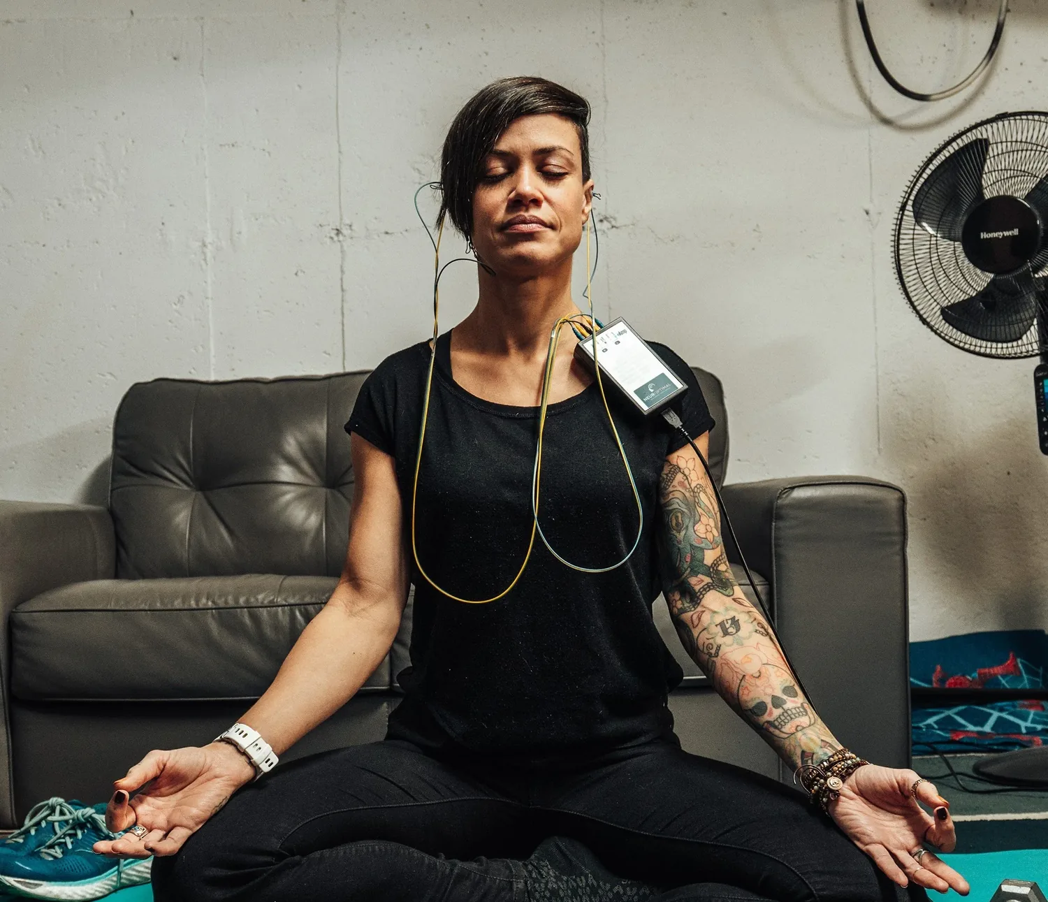 A woman with tattoos practicing yoga while receiving a neurofeedback session, sitting cross-legged with her hands resting on her knees, eyes closed, in a room with a gray couch and fans in the background.